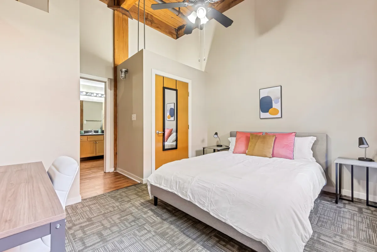 A spacious bedroom featuring a comfortable bed adorned with decorative pillows, exposed wooden ceiling beams adding warmth, a modern desk and chair providing a dedicated workspace, and tasteful artwork on the wall. Soft natural light streams in, creating a serene and inviting ambiance perfect for unwinding or getting some work done.