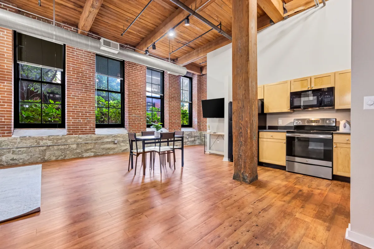 The image showcases a stylish apartment interior featuring exposed brick walls, impressive wooden beams, and expansive windows that flood the space with natural light. The open layout includes a modern kitchen area and a sleek dining table, evoking an atmosphere perfect for both relaxing evenings and lively gatherings.