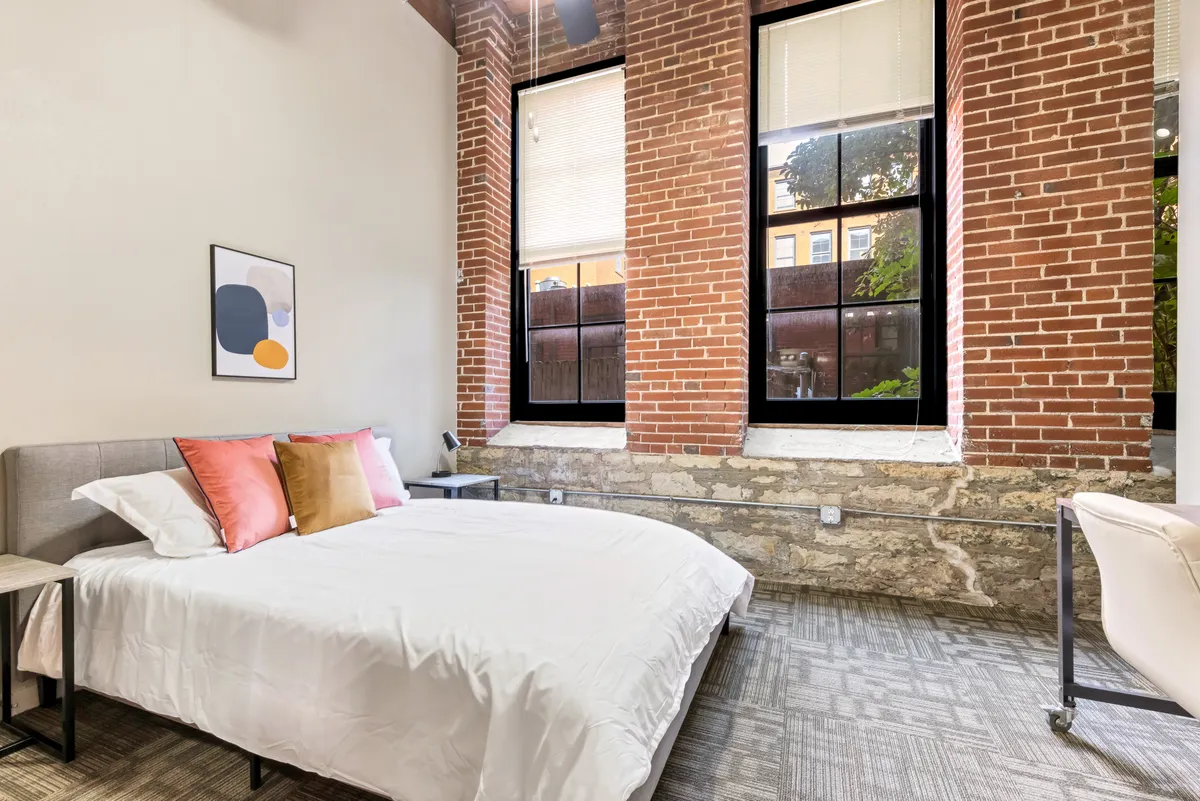 A modern, cozy bedroom flooded with natural light from expansive windows, featuring exposed brick walls, stylish artwork, and a comfortable bed. The inviting atmosphere suggests both relaxation and the potential for focused work, thanks to the abundance of daylight and open feel.