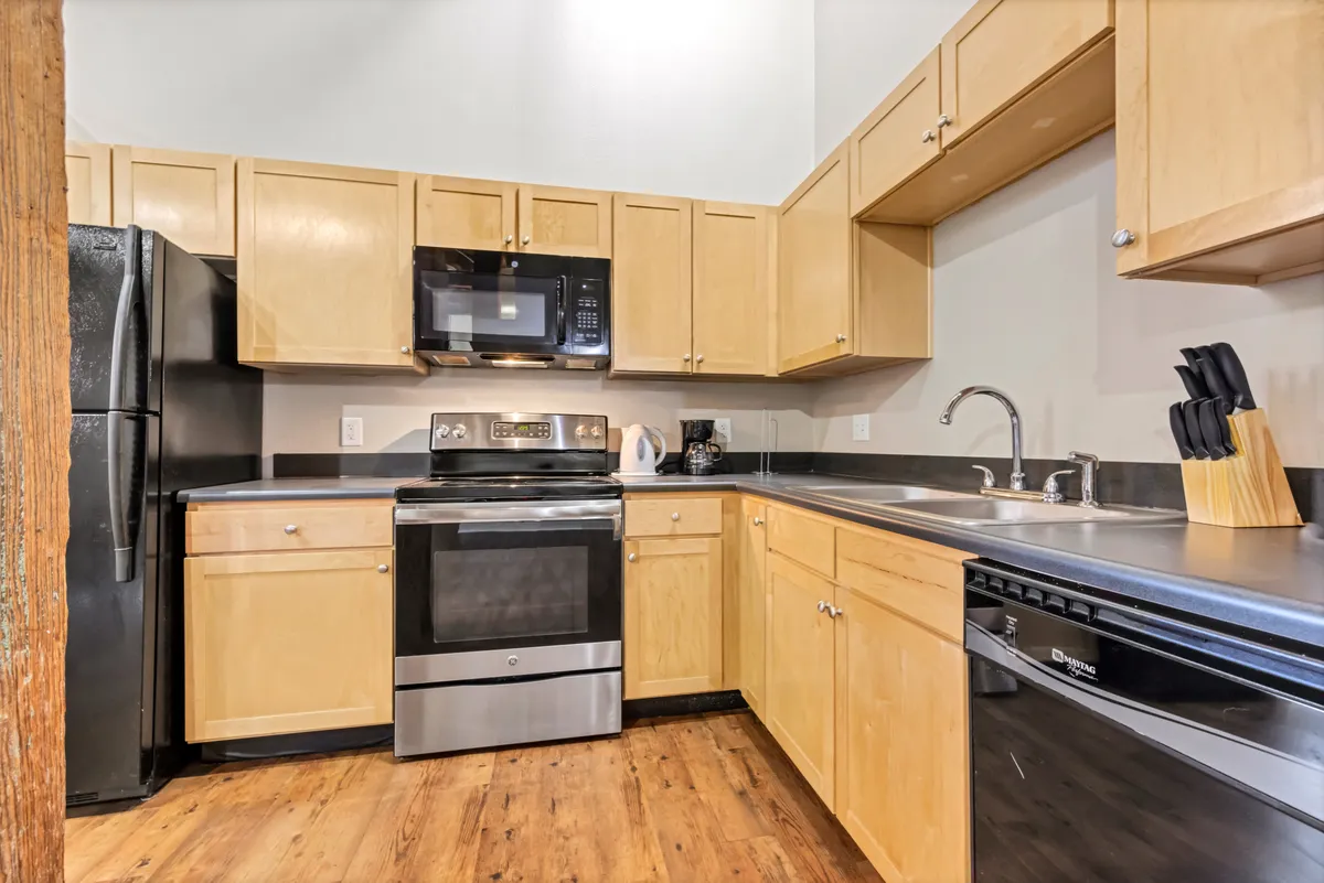 A modern kitchen interior featuring stainless steel appliances, wooden cabinetry, and dark countertops. The space is well-lit and arranged for both cooking and social gatherings, with ample workspace visible for meal preparation.