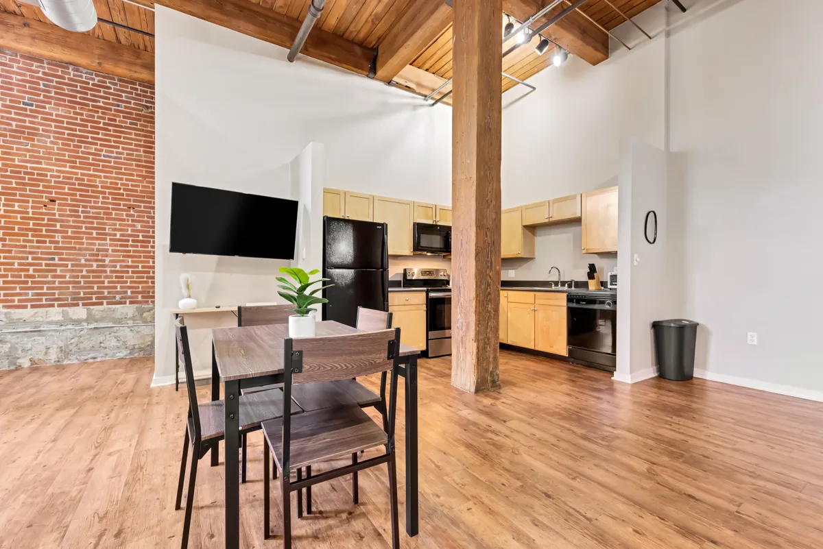 A contemporary open-plan living and dining space featuring a dining table set for a meal, a modern kitchen area with exposed brick wall and wooden beams, and a television. The arrangement encourages gatherings and conversation, with plenty of space for entertaining or relaxing.