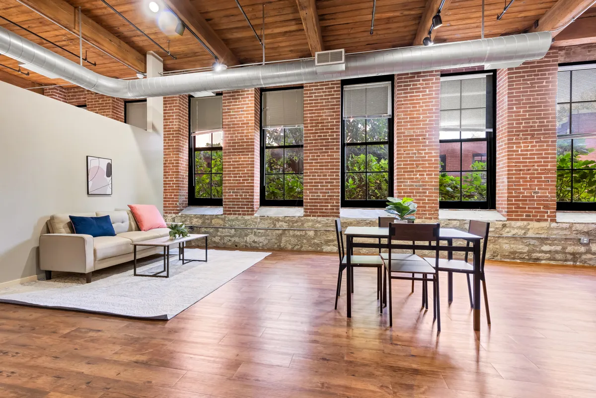 A warm and welcoming living area featuring a comfortable sofa, a dining table set for gatherings, expansive windows that flood the space with natural light, and classic exposed brick walls that evoke the building's historic charm.