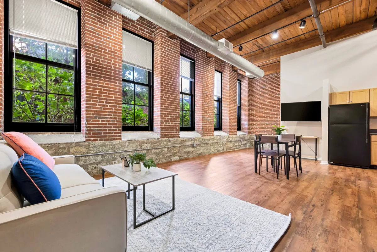 A modern living area with large windows letting in natural light, exposed brick walls, wooden ceiling, and thoughtfully arranged furniture. The space is designed for both relaxation and socializing, with views suggesting a connection to the lively neighborhood outside.